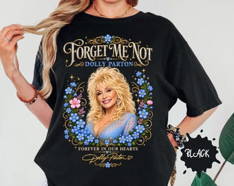 May include: A black t-shirt featuring a graphic of Dolly Parton surrounded by blue forget-me-not flowers and pink blossoms. The text reads "Forget Me Not", "Dolly Parton", and "Forever In Our Hearts" with a signature at the bottom.