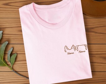 May include: A light pink short-sleeved t-shirt featuring a brown embroidered graphic on the chest. The design consists of two distinct dog ear outlines, each positioned above the word Name written in a cursive script font.