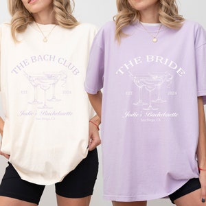 May include: Two oversized t-shirts with a bachelorette party theme. The ivory shirt says "The Bach Club" and the orchid shirt says "The Bride". Both shirts feature a graphic of three martini glasses and the text "Jodie's Bachelorette" with the location "San Diego, CA".