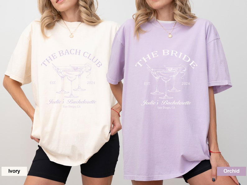 May include: Two oversized t-shirts with a bachelorette party theme. The ivory shirt says "The Bach Club" and the orchid shirt says "The Bride". Both shirts feature a graphic of three martini glasses and the text "Jodie's Bachelorette" with the location "San Diego, CA".