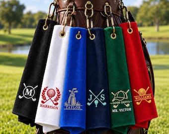 May include: Six rectangular golf towels in black, white, blue, navy, green, and red, each featuring gold-tone metal grommets and carabiner clips. Each towel displays unique embroidered golf-themed designs and names including M, Harrison, Taylor, J S, Mr. Victor, and Williams.