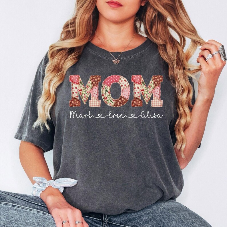 A woman with long, wavy blonde hair wearing a gray t-shirt with the word "MOM" printed on it.
