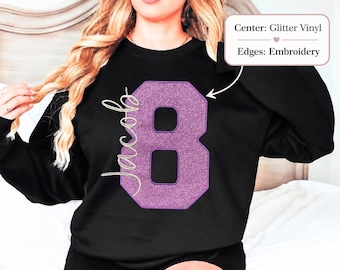 May include: A black long-sleeve sweatshirt featuring a large purple glitter vinyl number eight with embroidered edges. The name Jacob is written in white cursive script to the left of the number. A text box indicates Center: Glitter Vinyl and Edges: Embroidery.