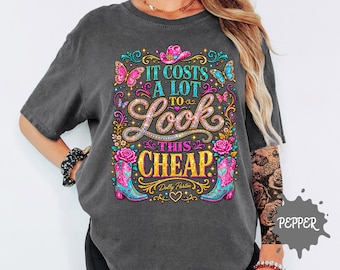 May include: A charcoal gray short-sleeve t-shirt featuring a colorful graphic print with the text "IT COSTS A LOT TO LOOK THIS CHEAP" and "Dolly Parton". The design includes pink cowboy boots, butterflies, roses, and a pink cowboy hat.