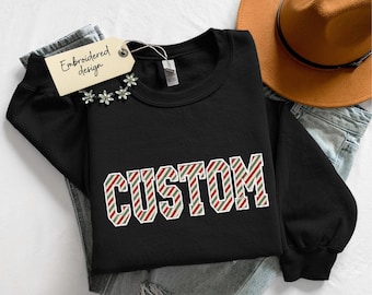 May include: A black long-sleeve sweatshirt featuring the word CUSTOM in a block font with a diagonal red, green, and white candy cane stripe pattern. A beige tag attached to the collar displays the handwritten text Embroidered design.