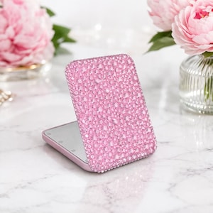 May include: A pink, rectangular compact mirror with a sparkling, crystal-covered exterior. The mirror is open, revealing a reflective surface. The background includes pink peonies in a glass vase, set on a marble surface.
