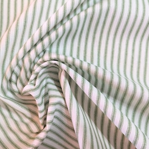 May include: Close-up of a white fabric with vertical green stripes. The fabric is folded and draped, creating shadows and texture. The stripes are evenly spaced and appear to be stitched. The overall effect is a classic, textured textile.