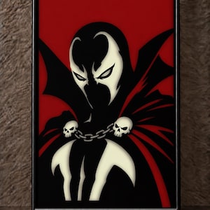 May include: A framed wall art piece featuring a stylized graphic illustration of a character with a black mask, white eye markings, and a flowing cape. The character wears a chain across the chest adorned with two white skull accents against a red background.