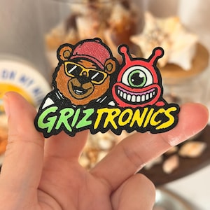 May include: A colorful patch featuring a cartoon bear wearing sunglasses and a red cap, alongside a red, one-eyed creature. The patch has the text "GRIZTRONICS" in green and yellow. The patch is held up by a hand.