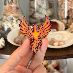 May include: A phoenix-shaped enamel pin featuring a vibrant gradient of red, orange, and yellow colors. The mythical bird is depicted with wings spread wide and detailed black outlines, showcasing a fiery design with a textured, colored surface.
