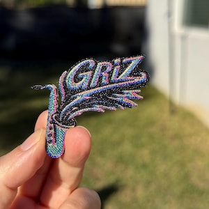 Op de afbeelding: Een zwarte geborduurde patch met een gestileerde saxofoon in iriserende blauwe, roze en groene draden. Het woord GRIZ is in een bijpassend iriserend lettertype boven de saxofoon geborduurd, met een spoor van kleurrijke, sprankelende borduurdraad.