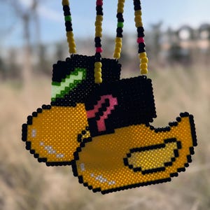 May include: Two pixelated yellow duck pendants with black accents and beaded necklaces. The ducks have black hats and green and pink details. The necklaces are yellow with black, green, and pink beads.