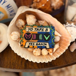 May include: A rectangular enamel pin shaped like a cassette tape with a split orange and blue background. The text YO TAPE B and YES GANJA is printed on the tape. Green vines wrap around the edges, and pink and green symbols are in the center.
