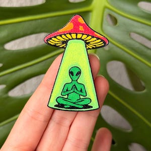 May include: A colorful patch featuring a meditating green alien under a red and yellow mushroom. The alien is in a lotus position, with hands together in prayer. The patch has a black border and is set against a green leaf.