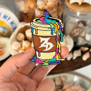 May include: A coffee cup pin featuring a tan cup with a brown sleeve and a white symbol. A vibrant, multi-colored liquid in shades of blue, green, yellow, orange, pink, and purple drips from the top and pools at the base of the cup.