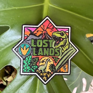 May include: A hexagonal embroidered patch featuring a vibrant landscape with a volcano, a green T-Rex, a long-necked dinosaur, and foliage. The text "LOST LANDS" is centered in black lettering, and a circular icon with a yellow dinosaur footprint is on the left.