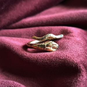 May include: A gold-toned metal ring shaped like a coiled snake. The ring features a detailed head with visible eyes and a textured, scaled body that wraps around to form an open, adjustable band design with a polished, reflective metallic finish.
