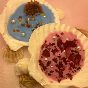 May include: Two seashell candles, one blue and one pink, adorned with dried flowers, petals, and faux pearls. The candles are set on a pink surface with additional seashells scattered around.
