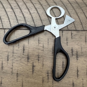 May include: A pair of stainless steel and black handled scissors, designed for cutting herbs. The scissors have a unique shape with a circular opening and a triangular blade. The handles are black and ergonomic.