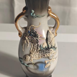 Könnte beinhalten: Eine kleine Keramikvase mit weißem Grund und dunkelgrauem Farbverlauf an Rand und Fuß. Sie besitzt zwei goldfarbene Henkel und ein erhabenes, strukturiertes Drachenrelief in Weiß, Schwarz und Blau, akzentuiert durch goldene Details.