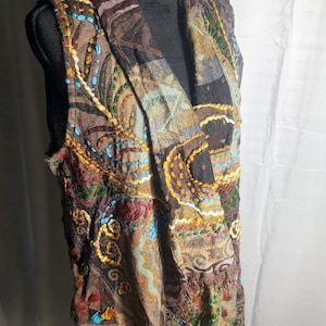 Puede incluir: Un chaleco sin mangas con una base de color marrón oscuro y bordados intrincados y multicolores en tonos amarillo, rojo, azul y verde. La prenda tiene un cuello ancho y drapeado, una superficie de tela texturizada y un dobladillo deshilachado.