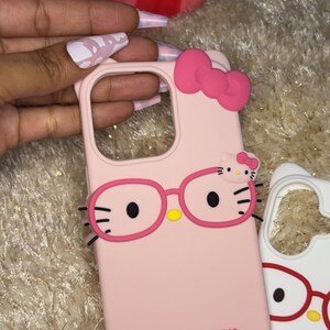 May include: Pink phone case featuring a Hello Kitty design with glasses and a bow. The case has the words "HELLO KITTY" printed on it. A second white phone case with a similar design is also visible.