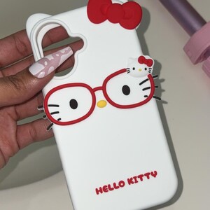 May include: White phone case featuring a Hello Kitty design. The case has a red bow, red glasses, and the words "HELLO KITTY" in red. The case is designed to fit a phone and has cutouts for the camera and buttons.