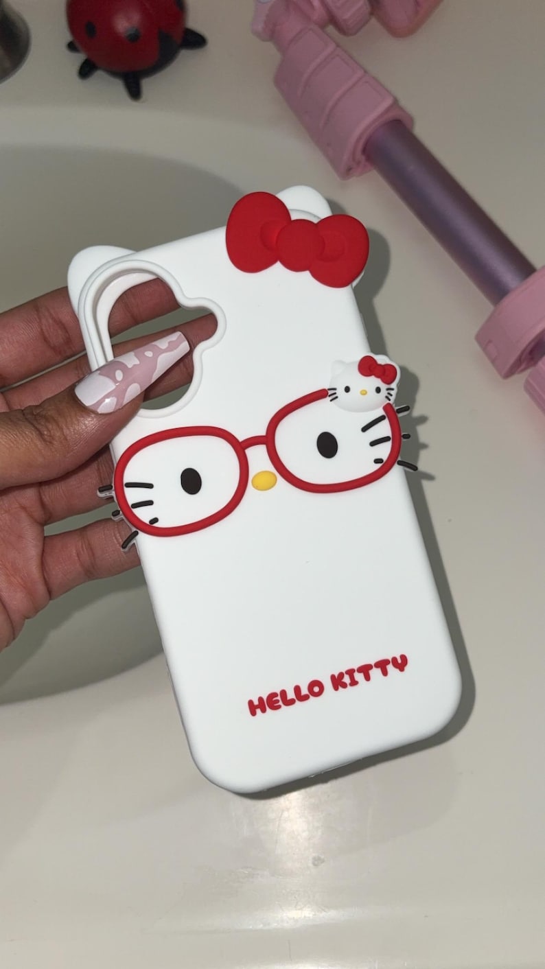 May include: White phone case featuring a Hello Kitty design. The case has a red bow, red glasses, and the words "HELLO KITTY" in red. The case is designed to fit a phone and has cutouts for the camera and buttons.