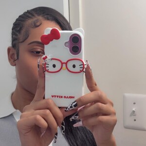 May include: White phone case featuring a Hello Kitty design with a red bow, red glasses, and the text "HELLO KILLA" in reverse. The case has a dual-camera cutout and a white background. The phone case is held up in front of a person.