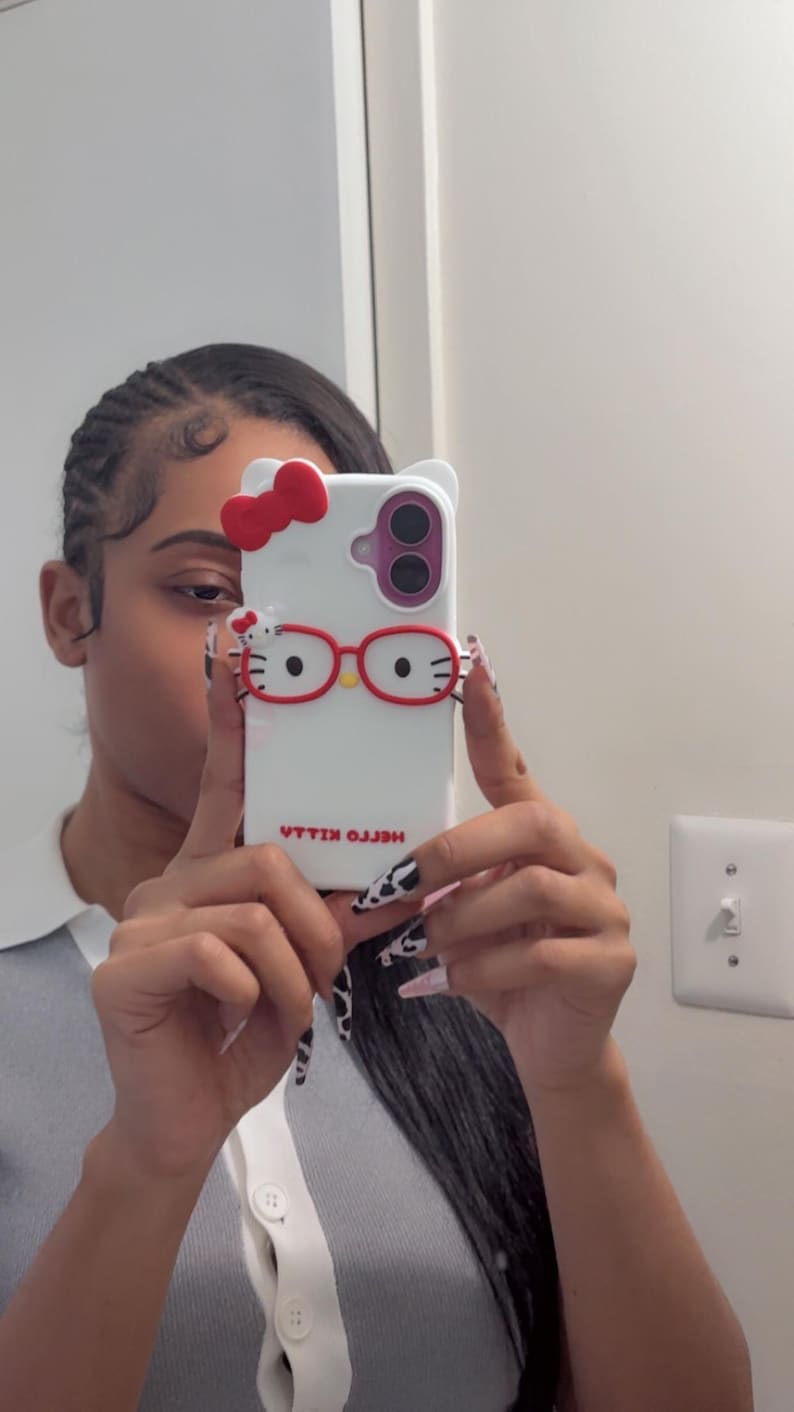 May include: White phone case featuring a Hello Kitty design with a red bow, red glasses, and the text "HELLO KILLA" in reverse. The case has a dual-camera cutout and a white background. The phone case is held up in front of a person.