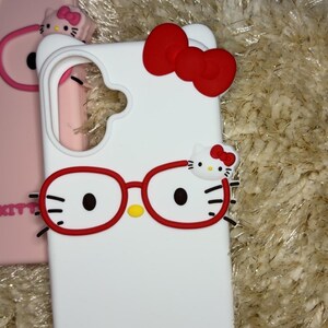 May include: White phone case featuring a red bow and Hello Kitty design. The case has red glasses and the words "HELLO KITTY" in red. Another pink phone case with a bow is visible in the background.