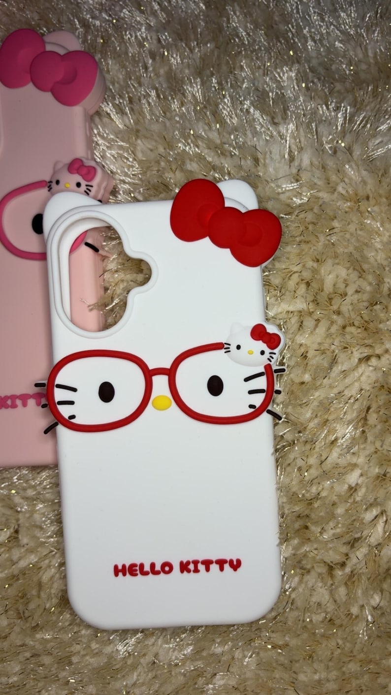 May include: White phone case featuring a red bow and Hello Kitty design. The case has red glasses and the words "HELLO KITTY" in red. Another pink phone case with a bow is visible in the background.