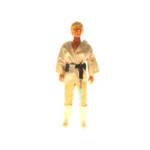 May include: A vintage action figure of Luke Skywalker from Star Wars. He is wearing a white robe with a black belt and holding a lightsaber. The figure has blonde hair and is standing on a white background.