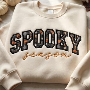 May include: A cream-coloured crewneck sweatshirt featuring the word SPOOKY in large, block-style letters filled with a dark pattern of orange pumpkins, white ghosts, and spiderwebs, with the word season embroidered in cursive orange thread below.
