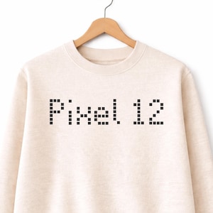 Könnte beinhalten: Beigefarbenes Sweatshirt mit dem Text "Pixel 12" in Pixel-Schrift. Das Sweatshirt hängt an einem Holzkleiderbügel vor weißem Hintergrund. Der Text ist schwarz.