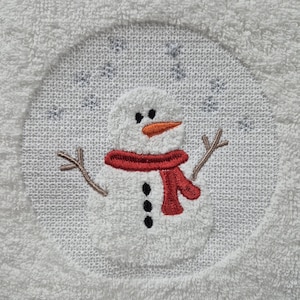 May include: White embroidered towel with a snowman design. The snowman is wearing a red scarf and has black eyes and buttons.