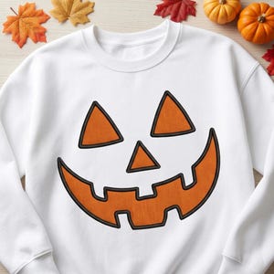May include: A white crewneck sweatshirt featuring a large, centered jack-o'-lantern face graphic. The design consists of two orange triangular eyes, a small orange triangular nose, and a wide, jagged orange mouth, all outlined in black.
