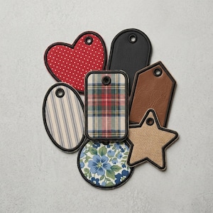 May include: A collection of seven assorted fabric and faux leather tags in various shapes including a heart, star, oval, and rectangles. The tags feature diverse patterns such as red polka dots, plaid, blue floral, stripes, solid black, and brown.