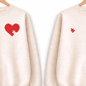 Op de afbeelding: Twee crèmekleurige sweatshirts. Eén heeft een rood hart met een ontbrekend puzzelstukje, de andere een rood puzzelstukje. Beide hangen aan houten hangers tegen een witte achtergrond. De sweatshirts zijn waarschijnlijk voor koppels of vrienden.