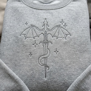 May include: A grey sweatshirt with an embroidered design of a sword with dragon wings and a serpent wrapped around it. The design is surrounded by four star-shaped accents. The sweatshirt is made of a soft, comfortable material.