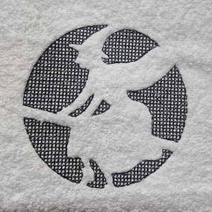 May include: White towel with a black witch silhouette design inside a broken circle. The witch is wearing a pointed hat and is riding a broom. The design is embroidered on the towel, which has a textured surface.