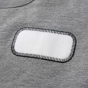 May include: A heather gray sweatshirt featuring a rectangular white fabric patch centered on the chest. The patch has rounded corners and is securely attached to the garment with thick, black blanket stitching along the entire outer edge.