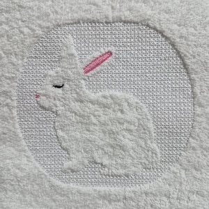 Può includere: Asciugamano bianco ricamato con un coniglio bianco con orecchie rosa. Il coniglio è sdraiato con gli occhi chiusi.