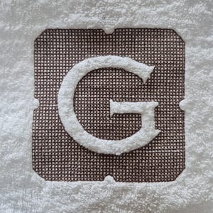 May include: White terry cloth towel with a large, embroidered letter "G". The letter is in a soft, white, raised texture, set against a brown, textured square. The towel has a white border.