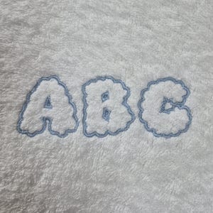 May include: White plush fabric with the letters "ABC" embroidered in a light blue, cloud-like font. The letters are puffy and have a soft, textured appearance, set against a fluffy white background.