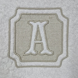 May include: White monogrammed towel with a large, embroidered letter "A" in a decorative frame. The frame and letter are in a light beige color, set against a white background. The towel has a soft, textured appearance.