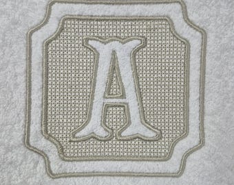 Könnte beinhalten: Weißes Handtuch mit einem gestickten Buchstaben "A" in einem dekorativen Rahmen. Der Buchstabe und der Rahmen sind in einem hellbeigen Farbton gehalten und heben sich vom weißen Hintergrund ab. Das Handtuch hat eine weiche, strukturierte Oberfläche.