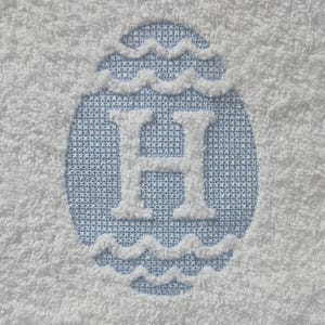May include: White terry cloth towel with a light blue oval design. The design features a white letter "H" in the center, surrounded by a wavy border. The background of the oval is a light blue fabric.