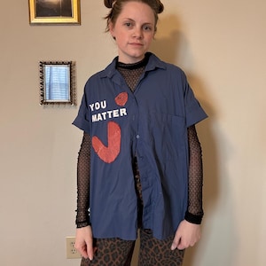 Könnte beinhalten: Ein marineblaues kurzärmeliges Hemd mit den Worten "YOU MATTER" und einem roten herzförmigen Design. Das Hemd wird über einem schwarzen Mesh-Top und einer Leopardenhose getragen.