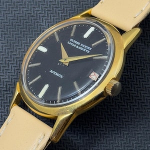 May include: A gold-tone wristwatch with a black face and white markings. The watch face reads "ULYSSE NARDIN LOCLE & GENEVE" and "AUTOMATIC".